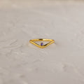 The Amethyst Drop Curve Ring features a delicate gold band with an amethyst accent, showcased on a textured white surface.