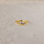 The Amethyst Drop Curve Ring features a delicate gold band with an amethyst accent, showcased on a textured white surface.