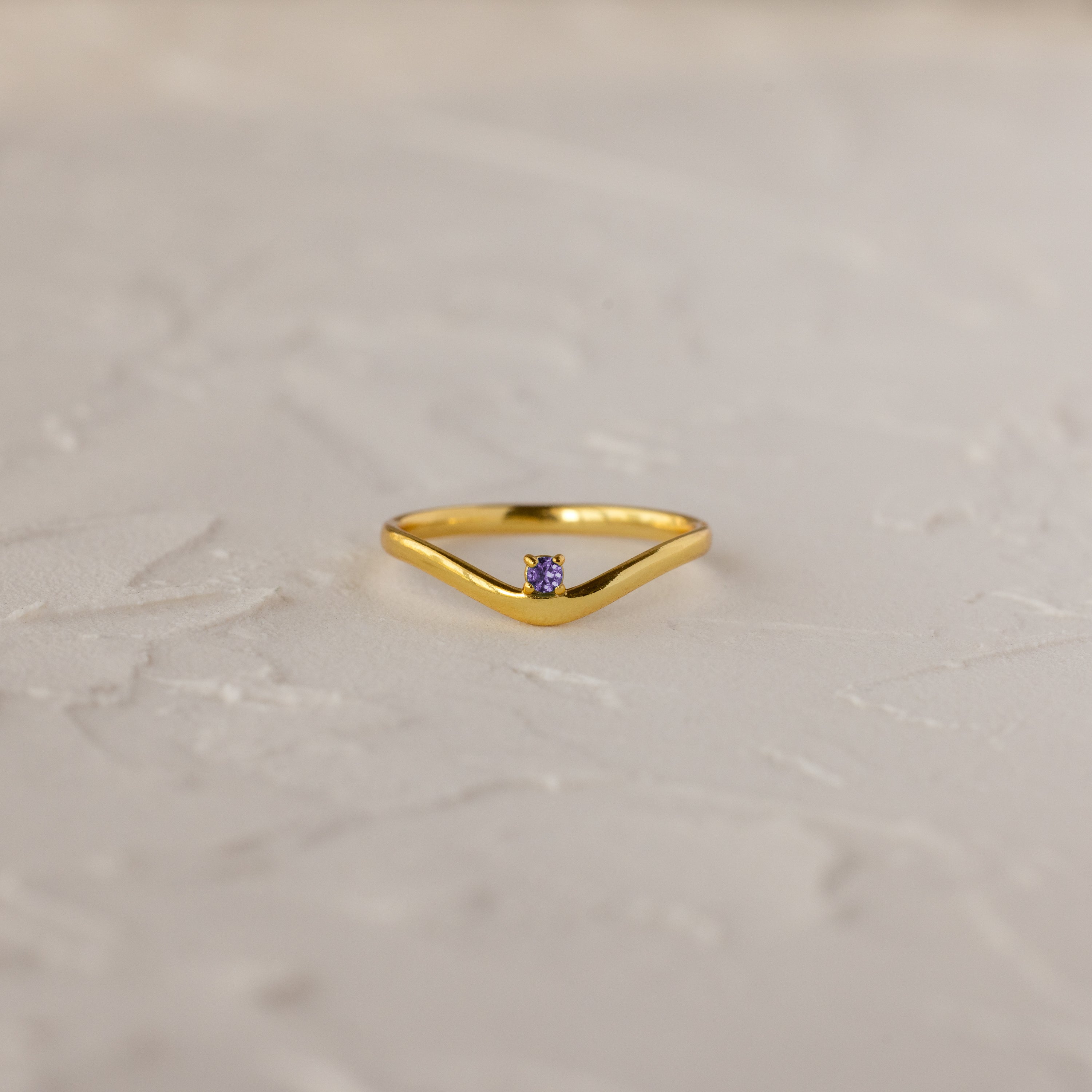 The Amethyst Drop Curve Ring features a delicate gold band with an amethyst accent, showcased on a textured white surface.