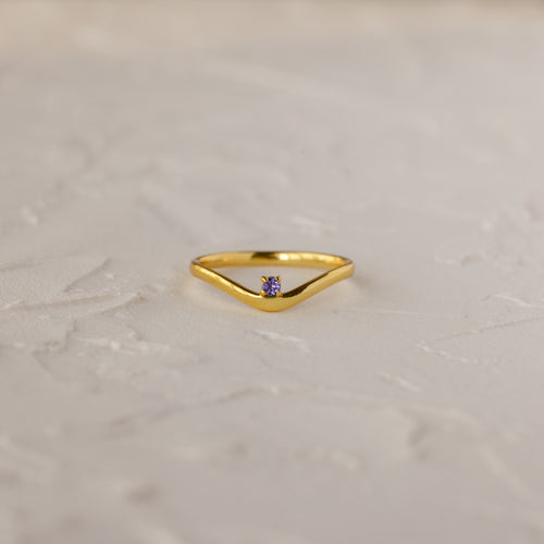 The Amethyst Drop Curve Ring features a delicate gold band with an amethyst accent, showcased on a textured white surface.