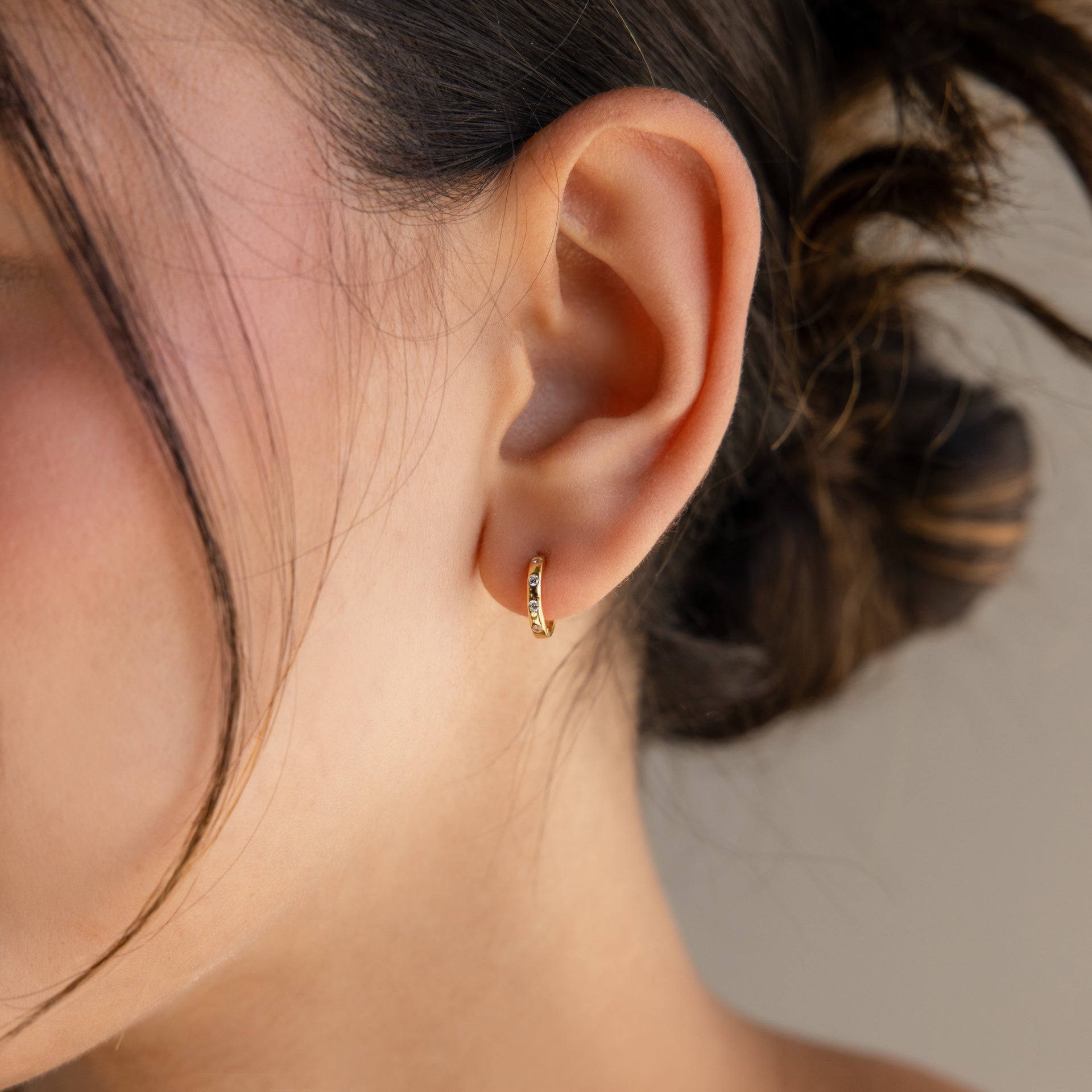 A womans ear with hair tied back showcases the Mini Diamond Sprinkle Huggies—small gold hoops that sparkle like diamonds for a delicate, elegant finish.