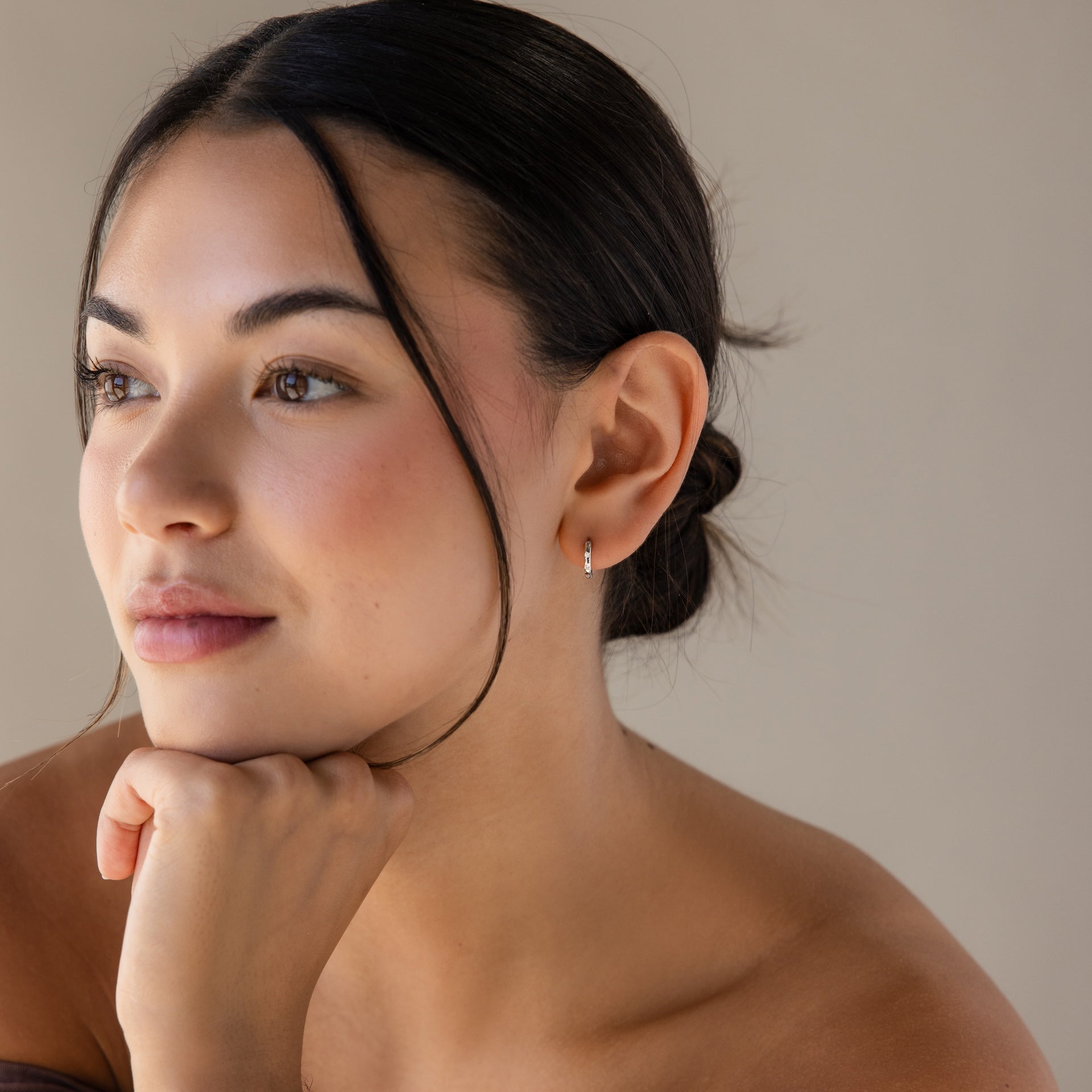 A woman with dark hair in a low bun rests her chin on her hand, gazing thoughtfully to the side while wearing Mini Diamond Sprinkle Huggies in Silver that add a hint of sparkle to her look.