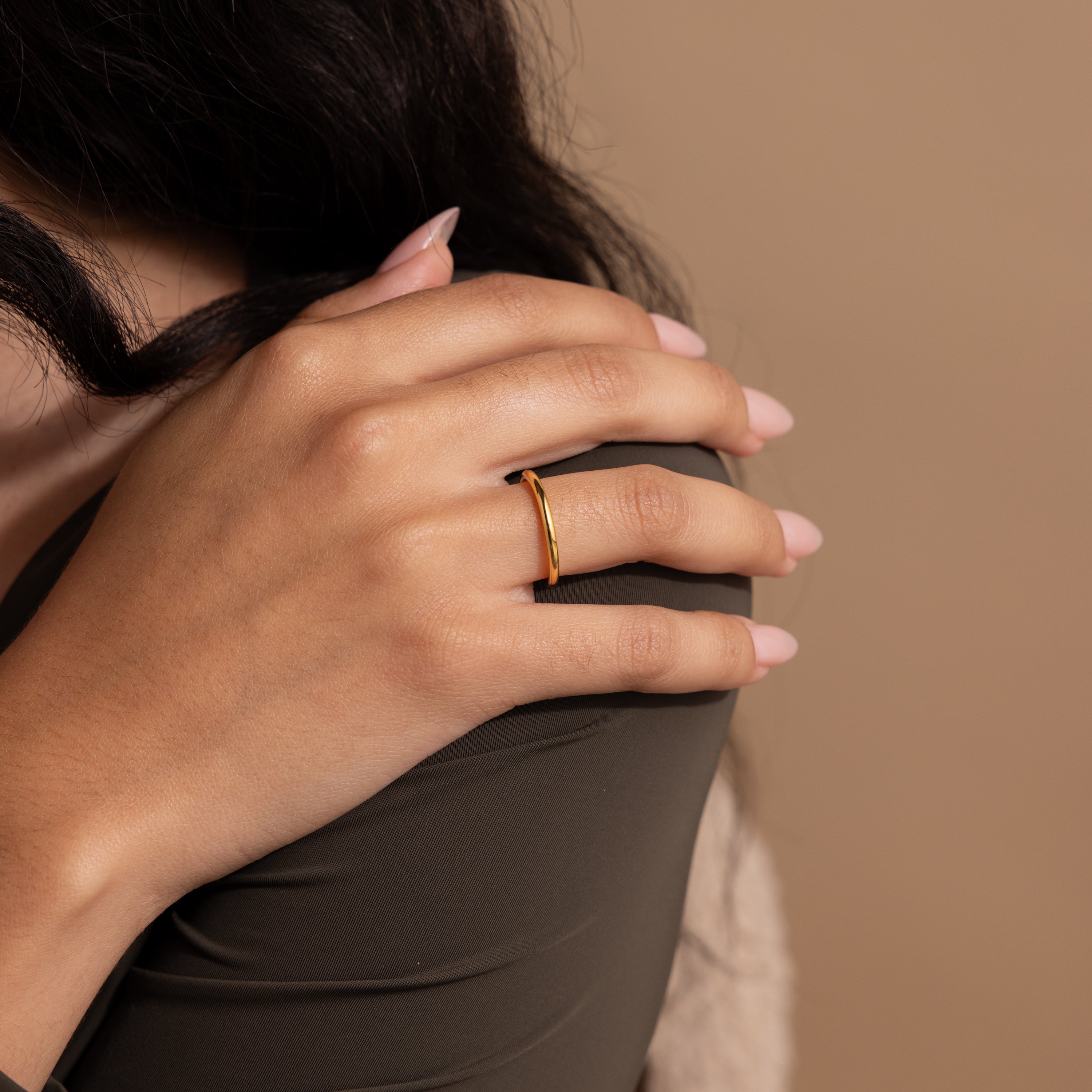 A hand with long, pale pink nails wears a dainty ring and the Minimal Stacking Ring in 18K Gold against a beige background.