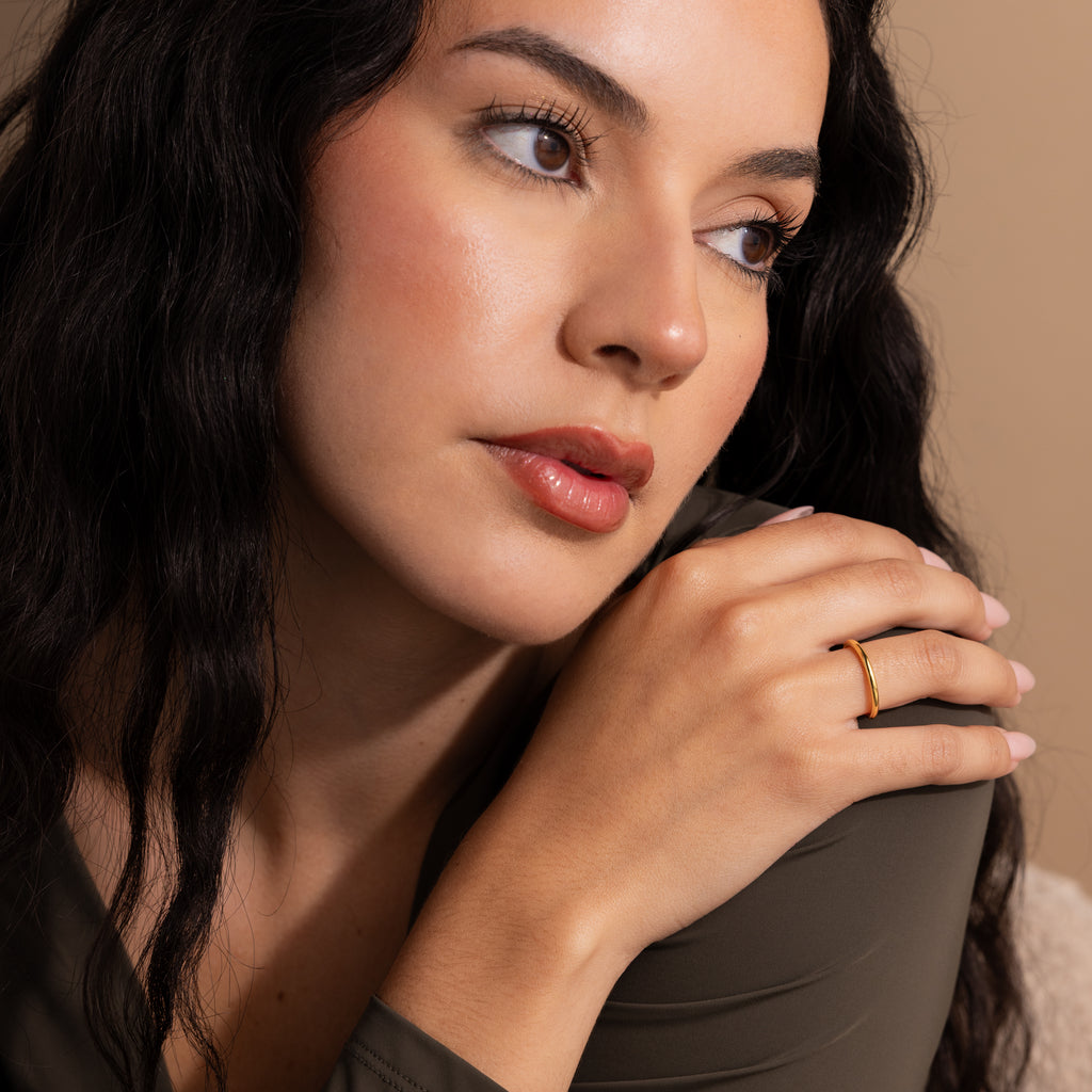 A woman with long dark hair wears the Minimal Stacking Ring in 18K Gold, looking thoughtfully to the side against a beige background.