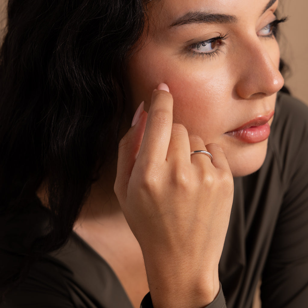 Woman with long dark hair touches her cheek while wearing the Minimal Stacking Ring in Silver and an olive green top, looking to the side.