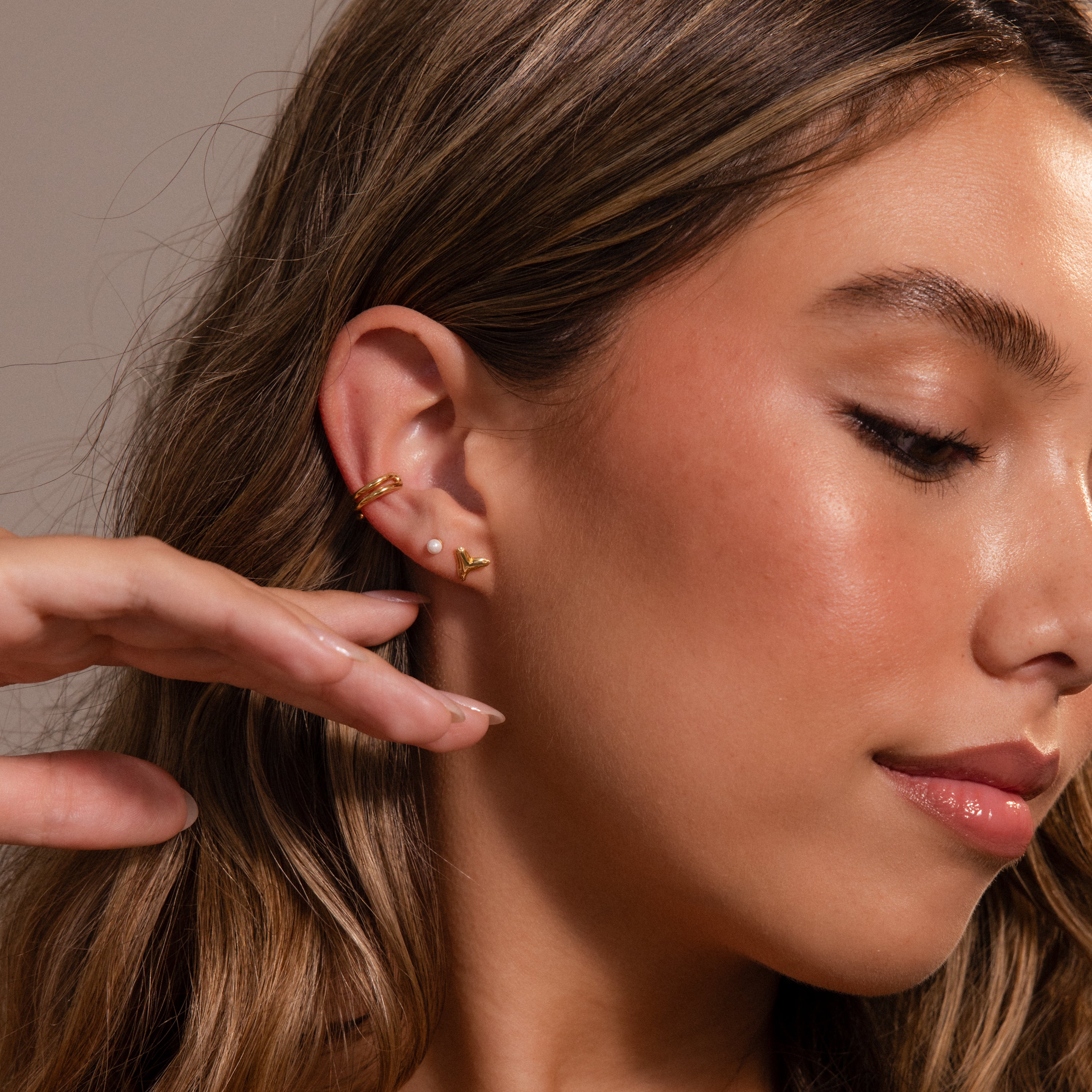 Woman with long brown hair wearing gold ear cuffs, a small pearl earring, and Shark Tooth Flatback Studs—a chic beach accessory—hand gracefully raised near her face.