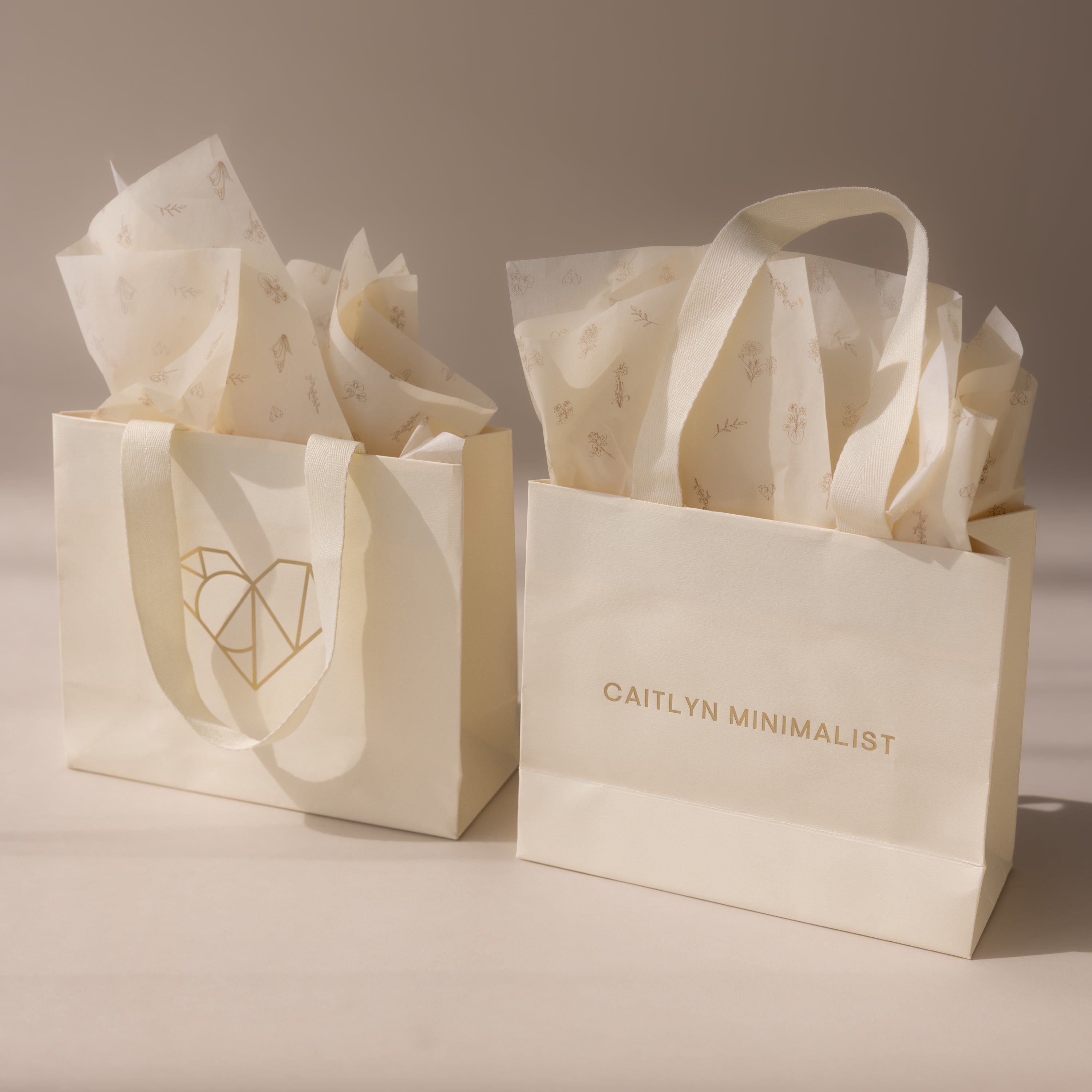 The Gift Bag Add-On includes two cream gift bags with tissue paper, featuring minimalist gold designs and branding—an elegant way to add a special finishing touch to any present.
