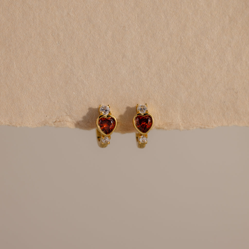 Garnet Heart Hoops featuring red heart-shaped stones and small clear accents, resembling diamond huggies, displayed on a textured beige surface.