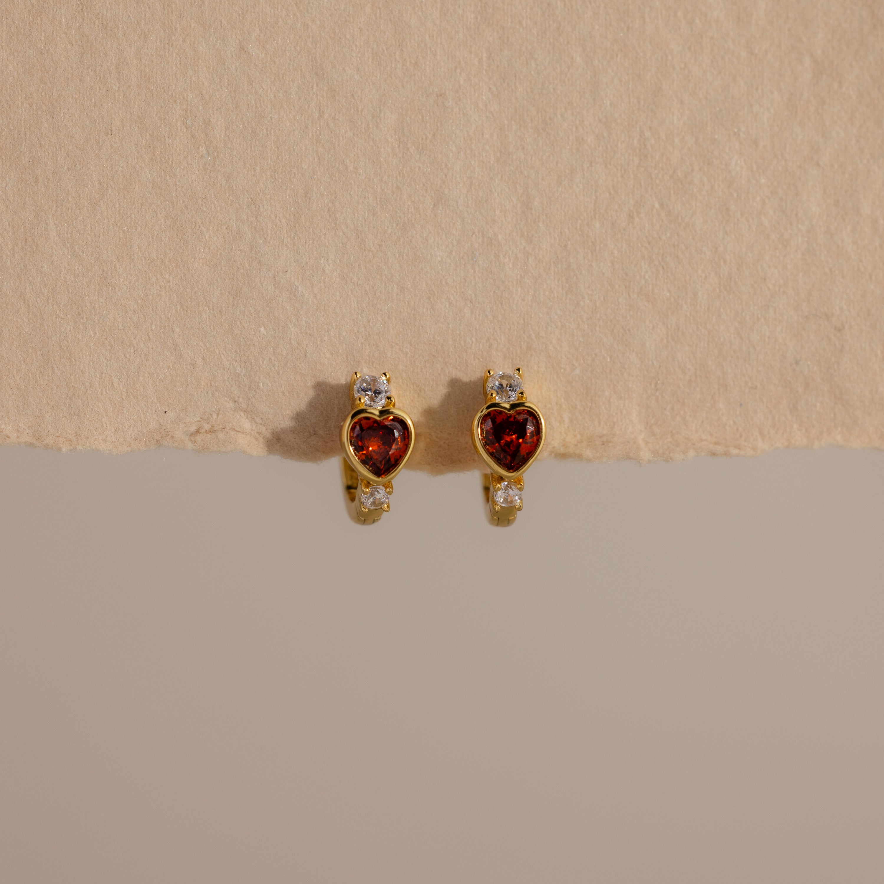 Garnet Heart Hoops featuring red heart-shaped stones and small clear accents, resembling diamond huggies, displayed on a textured beige surface.