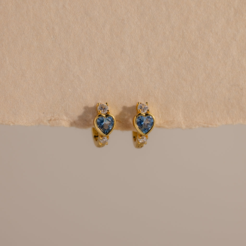 Aquamarine Heart Hoops: Gold earrings with blue heart-shaped stones and small clear gems, displayed on a textured beige surface—perfect as elegant birthstone earrings.