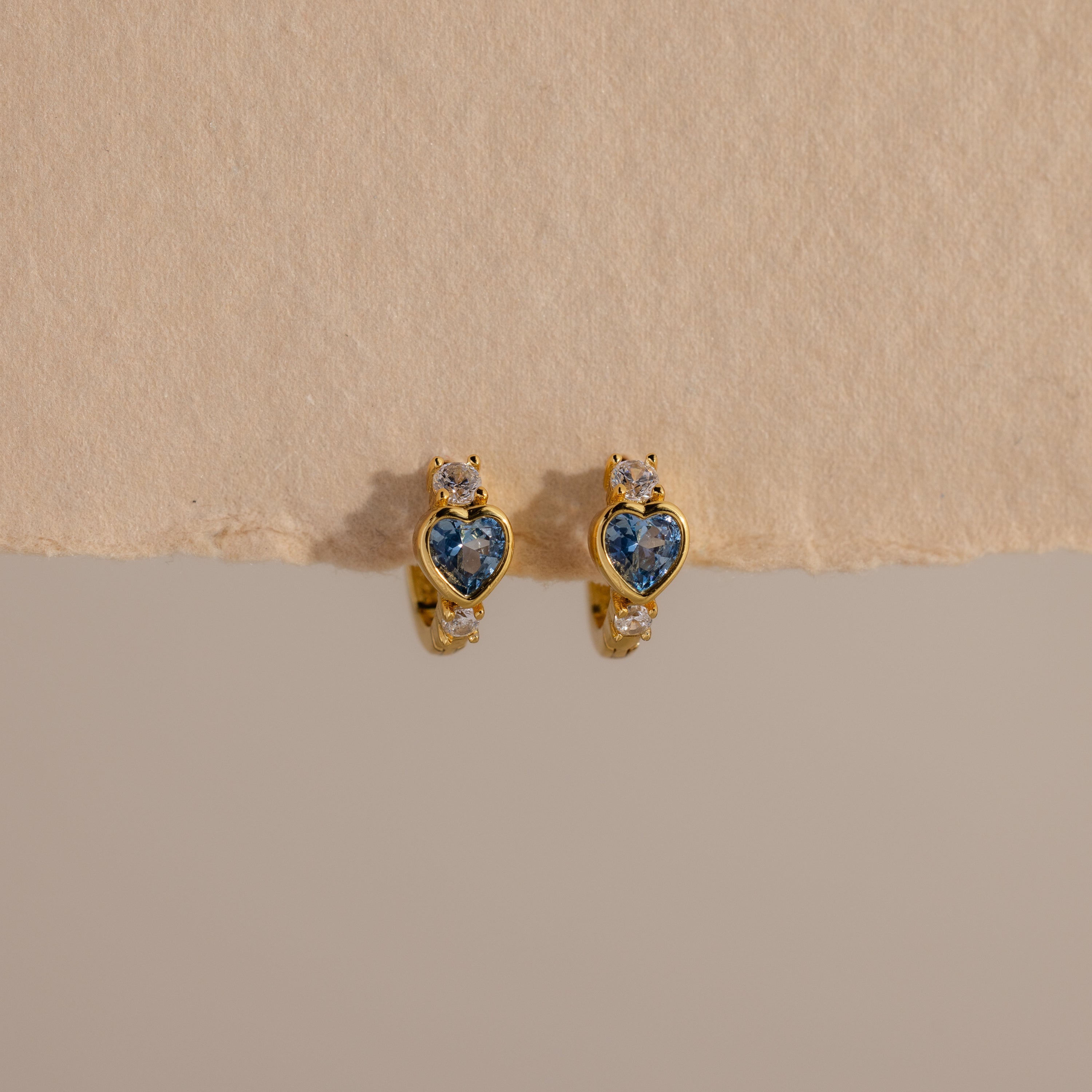 Aquamarine Heart Hoops: Gold earrings with blue heart-shaped stones and small clear gems, displayed on a textured beige surface—perfect as elegant birthstone earrings.