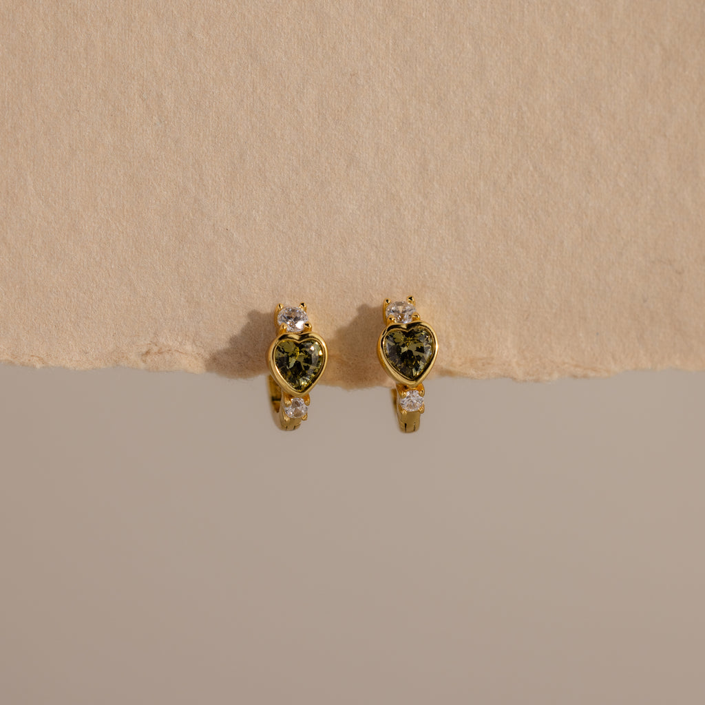 Peridot Heart Hoops featuring heart-shaped green stones and small clear accents, displayed on a beige surface—ideal as birthstone earrings or stylish gemstone huggies for any occasion.