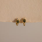 Peridot Heart Hoops featuring heart-shaped green stones and small clear accents, displayed on a beige surface—ideal as birthstone earrings or stylish gemstone huggies for any occasion.