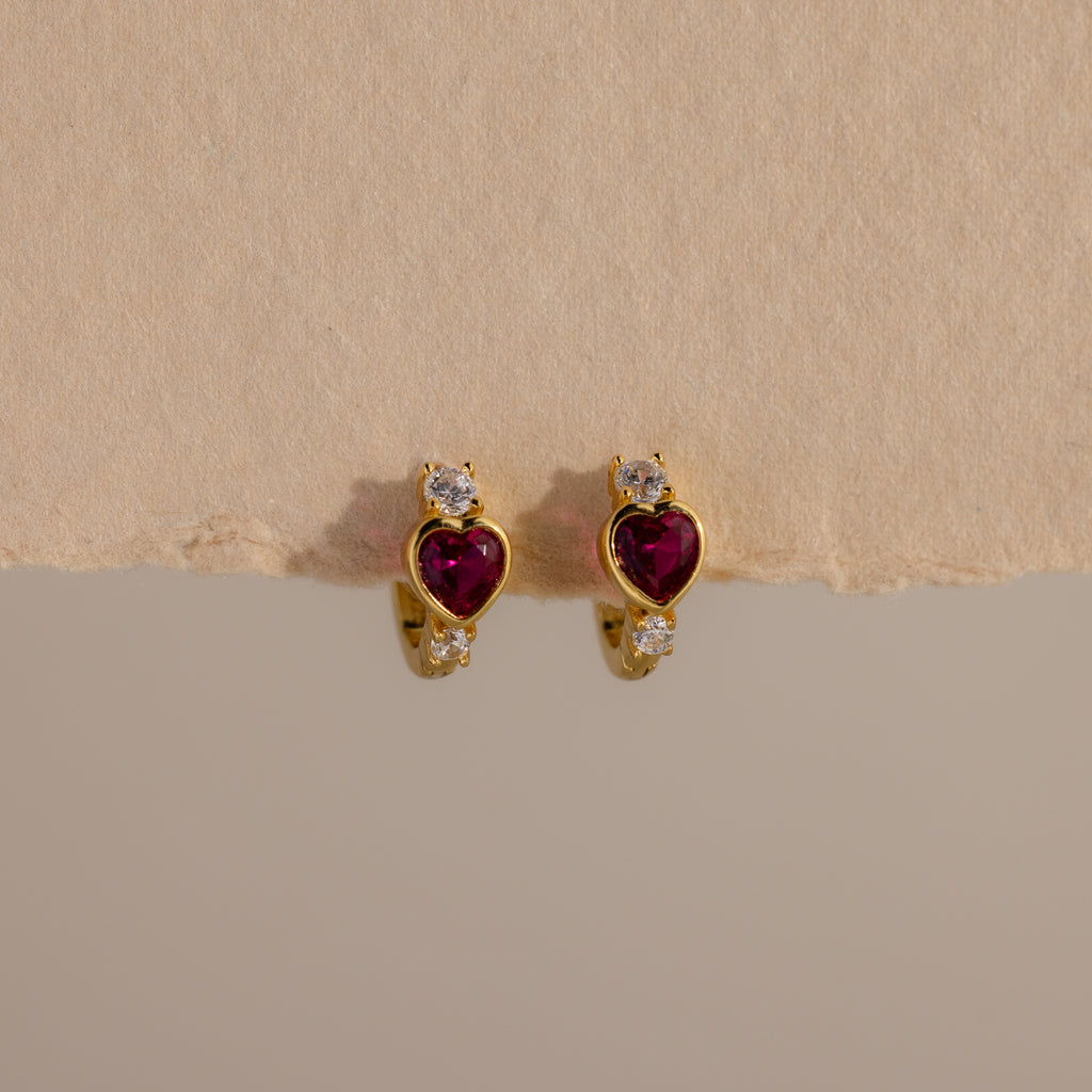 Ruby Heart Hoops: Gold earrings with heart-shaped red stones and small white gems, displayed on a textured beige background—perfect as dazzling birthstone earrings.