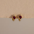 Ruby Heart Hoops: Gold earrings with heart-shaped red stones and small white gems, displayed on a textured beige background—perfect as dazzling birthstone earrings.