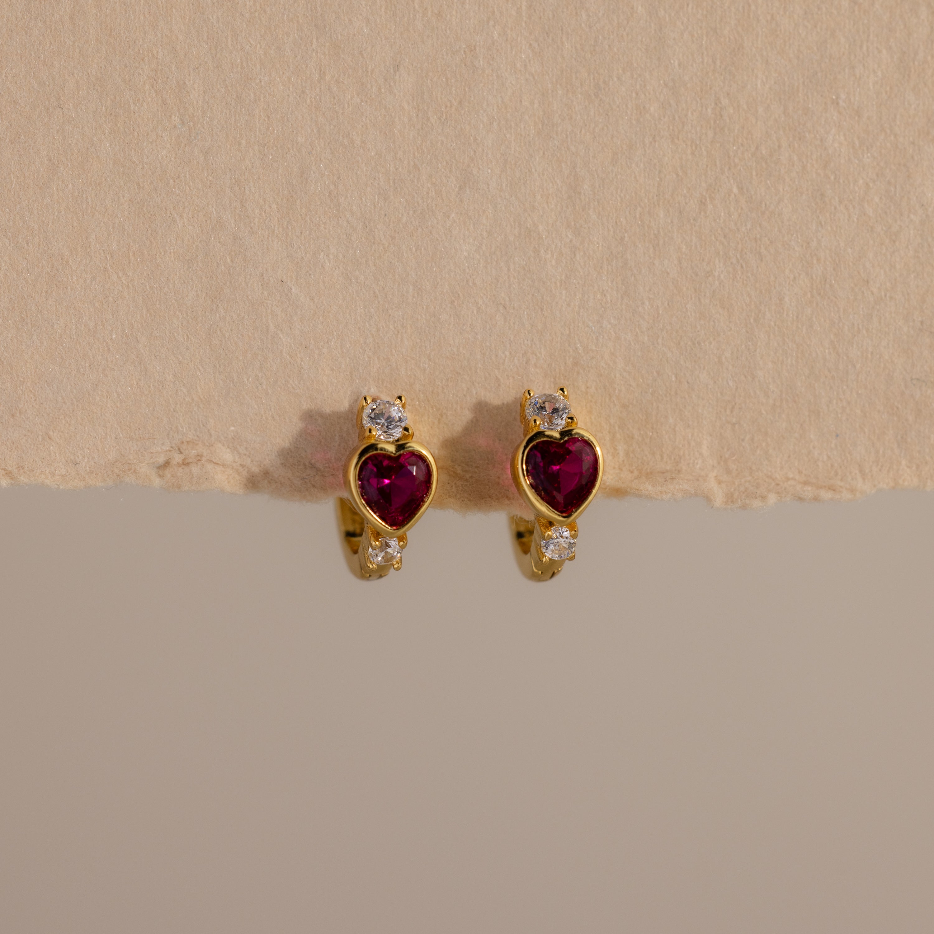 Ruby Heart Hoops: Gold earrings with heart-shaped red stones and small white gems, displayed on a textured beige background—perfect as dazzling birthstone earrings.