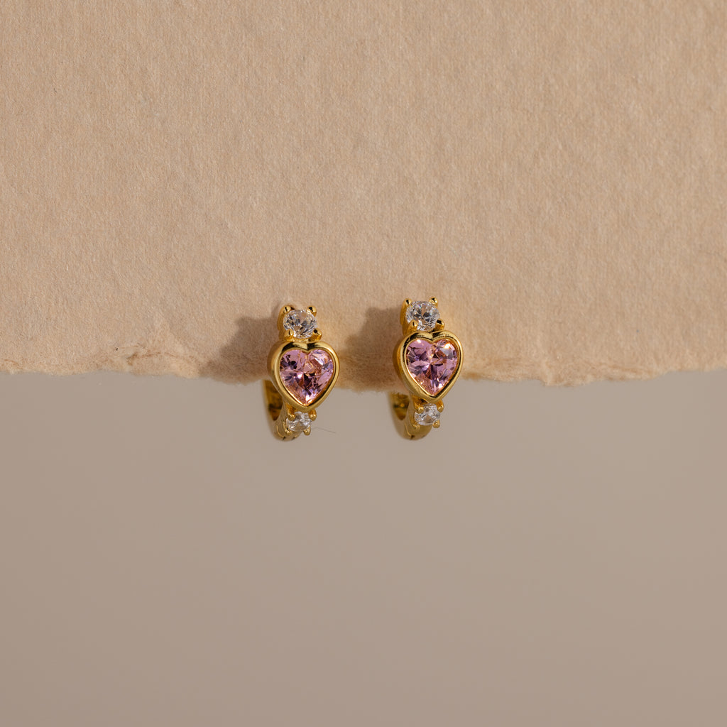 Pink Tourmaline Heart Hoops feature gold earrings with pink heart-shaped stones and clear round accents, showcased on a neutral textured background.