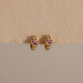 Pink Tourmaline Heart Hoops feature gold earrings with pink heart-shaped stones and clear round accents, showcased on a neutral textured background.