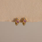 Pink Tourmaline Heart Hoops feature gold earrings with pink heart-shaped stones and clear round accents, showcased on a neutral textured background.
