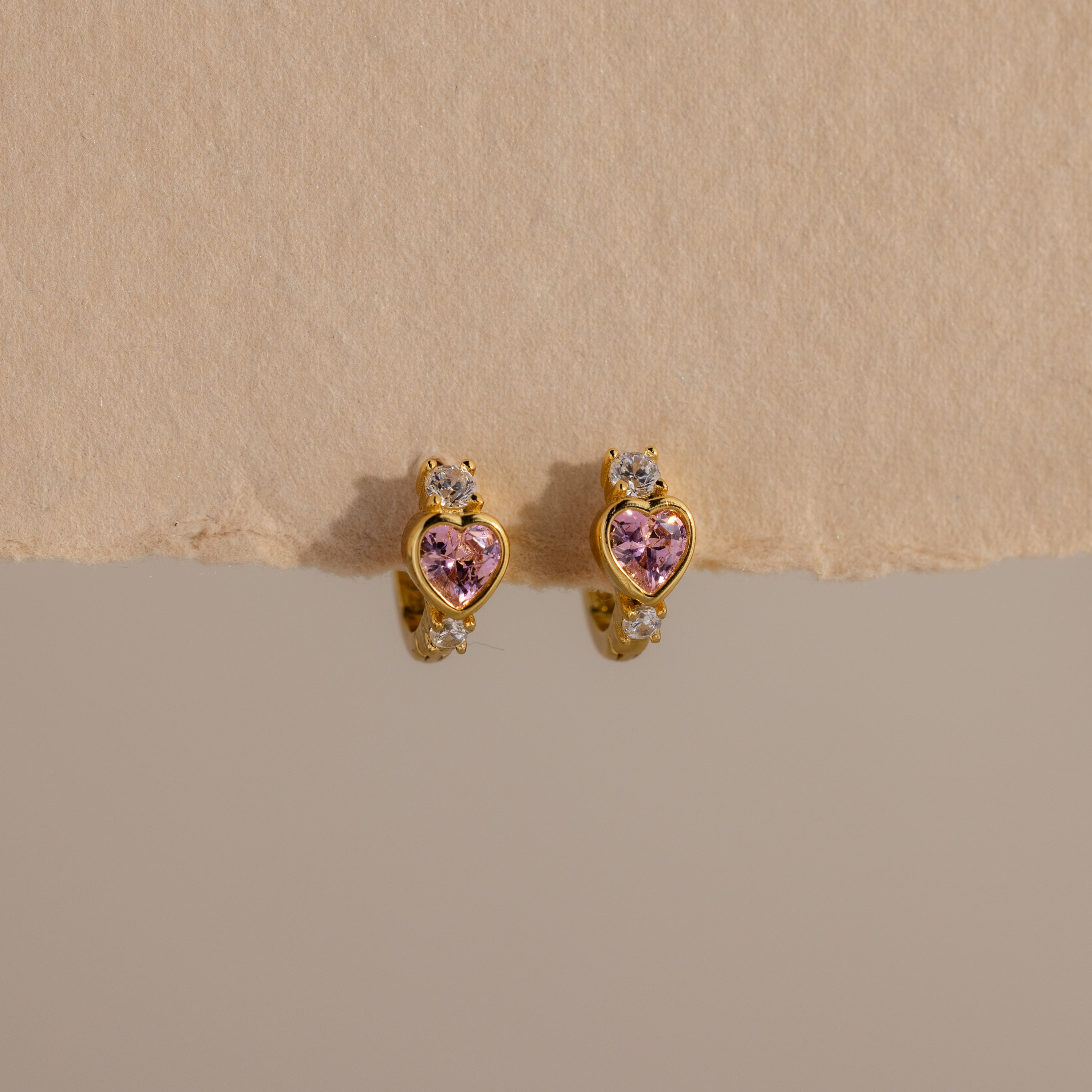 Pink Tourmaline Heart Hoops feature gold earrings with pink heart-shaped stones and clear round accents, showcased on a neutral textured background.