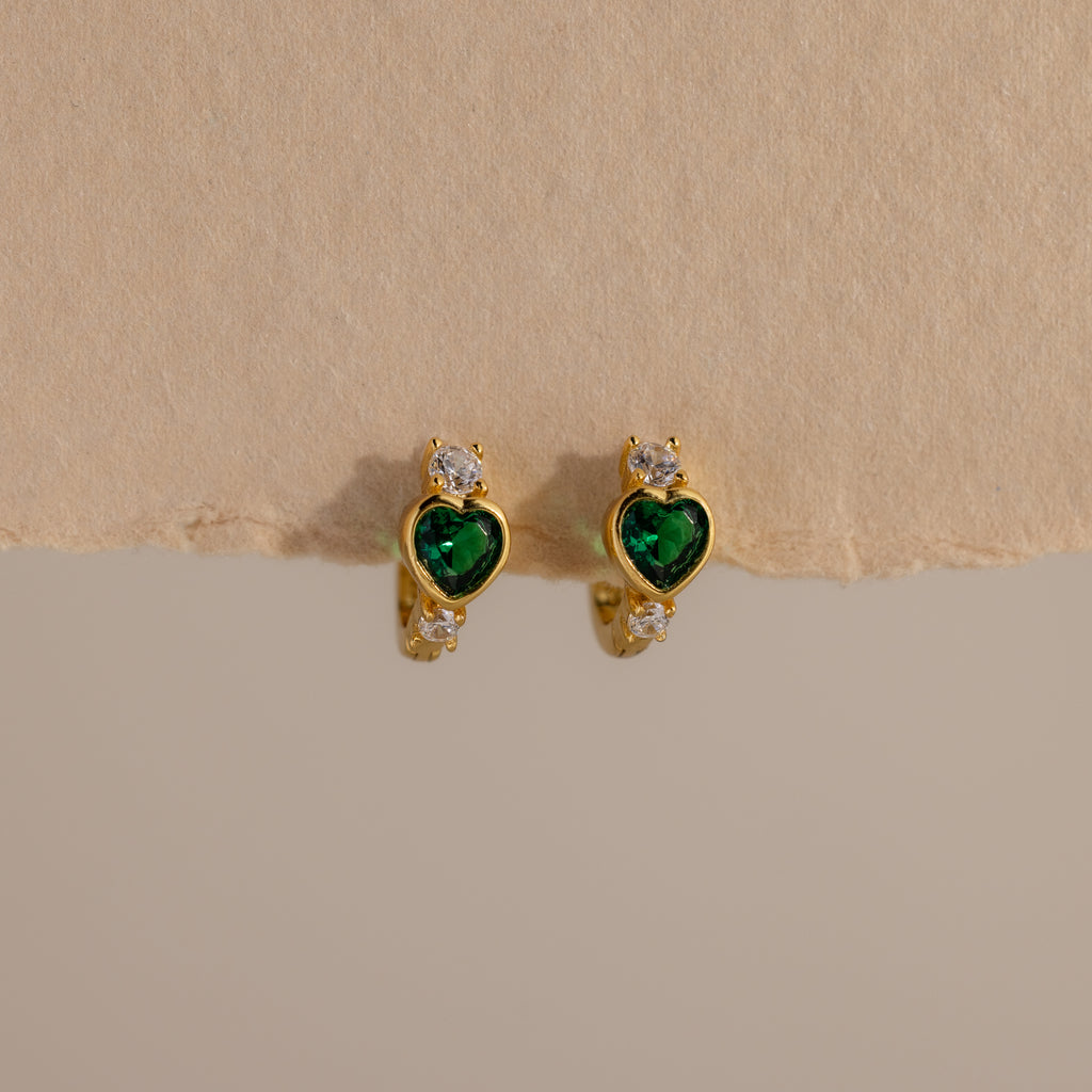 Emerald Heart Hoops feature green heart-shaped gems and small white stones, designed to add charm to any look—gemstone huggies displayed on a beige background.