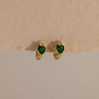 Emerald Heart Hoops feature green heart-shaped gems and small white stones, designed to add charm to any look—gemstone huggies displayed on a beige background.