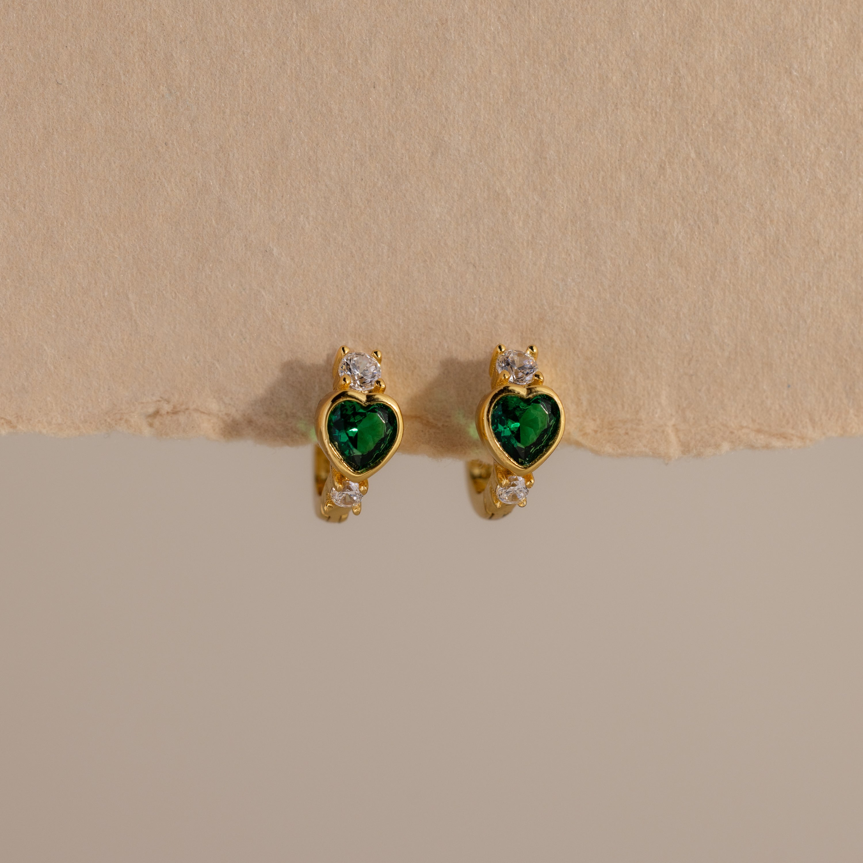 Emerald Heart Hoops feature green heart-shaped gems and small white stones, designed to add charm to any look—gemstone huggies displayed on a beige background.