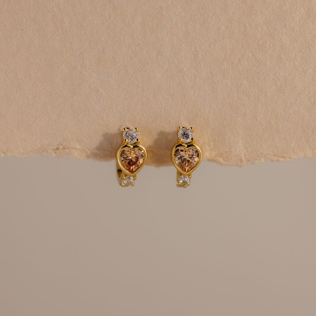 Topaz Heart Hoops are birthstone earrings featuring heart-shaped topaz gems with small clear stones, shown on a textured beige background.