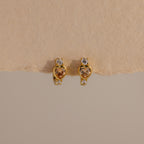 Topaz Heart Hoops are birthstone earrings featuring heart-shaped topaz gems with small clear stones, shown on a textured beige background.