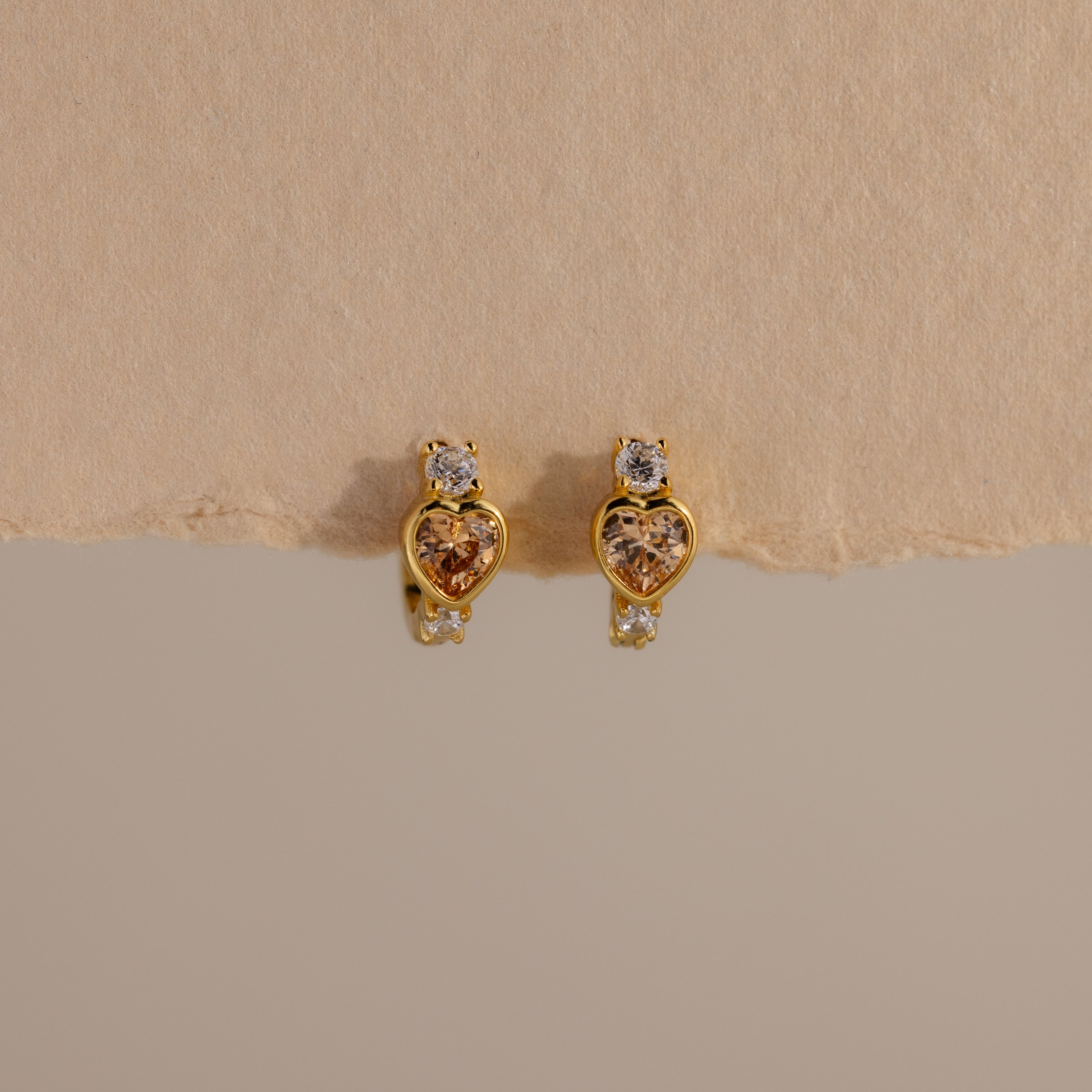 Topaz Heart Hoops are birthstone earrings featuring heart-shaped topaz gems with small clear stones, shown on a textured beige background.