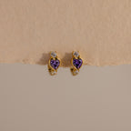 Amethyst Heart Hoops featuring purple heart-shaped stones and small clear gems, reminiscent of diamond huggies, displayed on a beige textured surface.