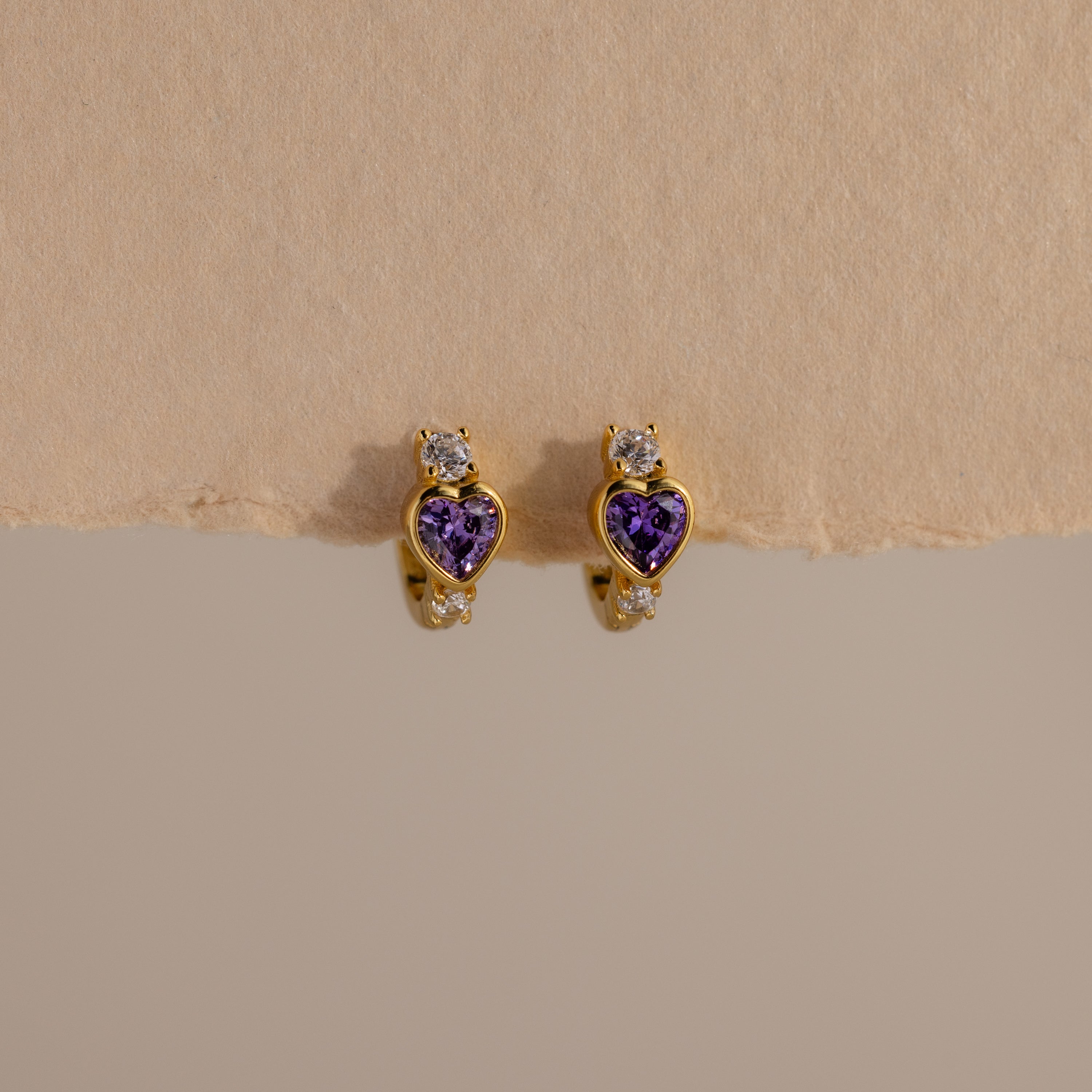 Amethyst Heart Hoops featuring purple heart-shaped stones and small clear gems, reminiscent of diamond huggies, displayed on a beige textured surface.