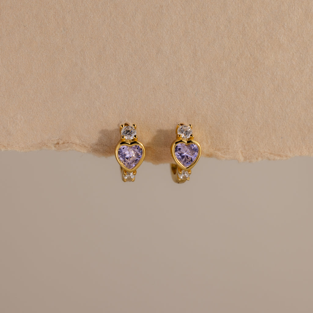 The Alexandrite Heart Hoops feature gold tones, purple heart-shaped stones, and small clear gems, capturing the charm of birthstone earrings. Their sparkle stands out beautifully against a beige background.