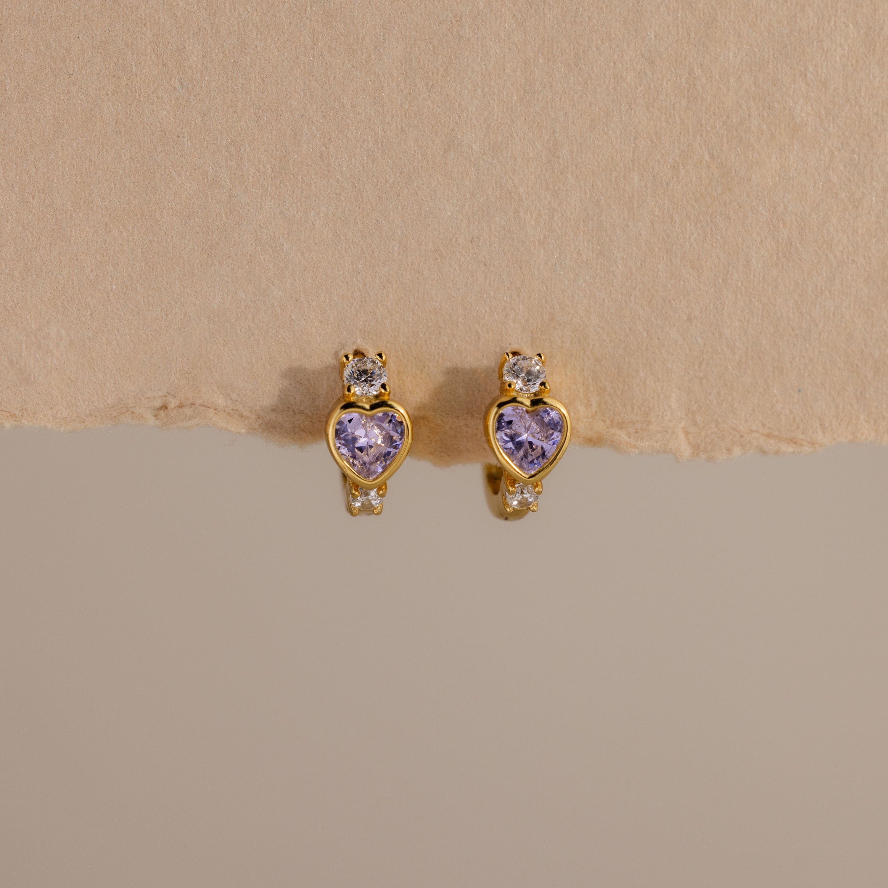 The Alexandrite Heart Hoops feature gold tones, purple heart-shaped stones, and small clear gems, capturing the charm of birthstone earrings. Their sparkle stands out beautifully against a beige background.