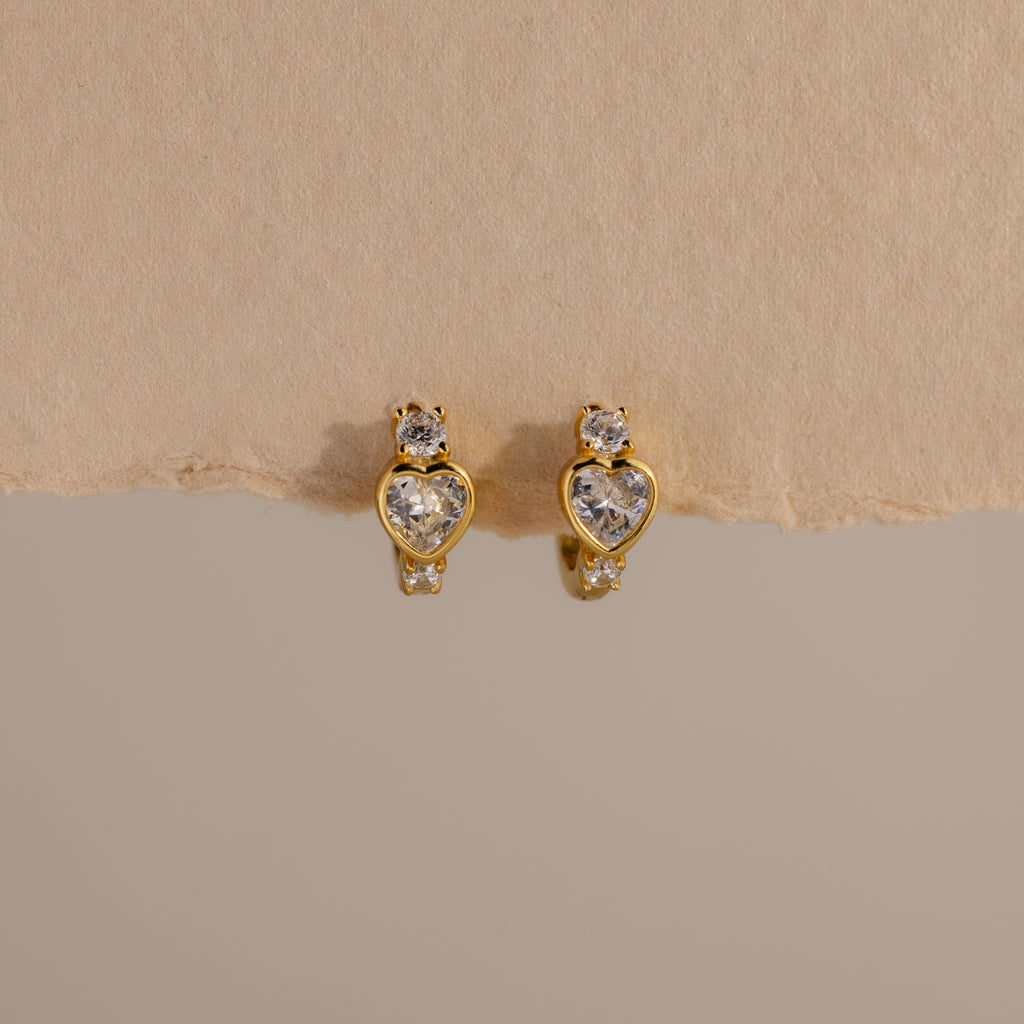 Diamond Heart Hoops featuring heart-shaped and round clear gemstones are elegantly displayed on a beige textured background.