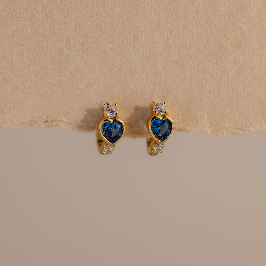 Blue Zircon Heart Hoops with sparkling clear stones are displayed on a beige textured background. These earrings add unique charm to any jewelry collection.