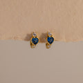 Blue Zircon Heart Hoops with sparkling clear stones are displayed on a beige textured background. These earrings add unique charm to any jewelry collection.