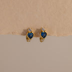 Blue Zircon Heart Hoops with sparkling clear stones are displayed on a beige textured background. These earrings add unique charm to any jewelry collection.