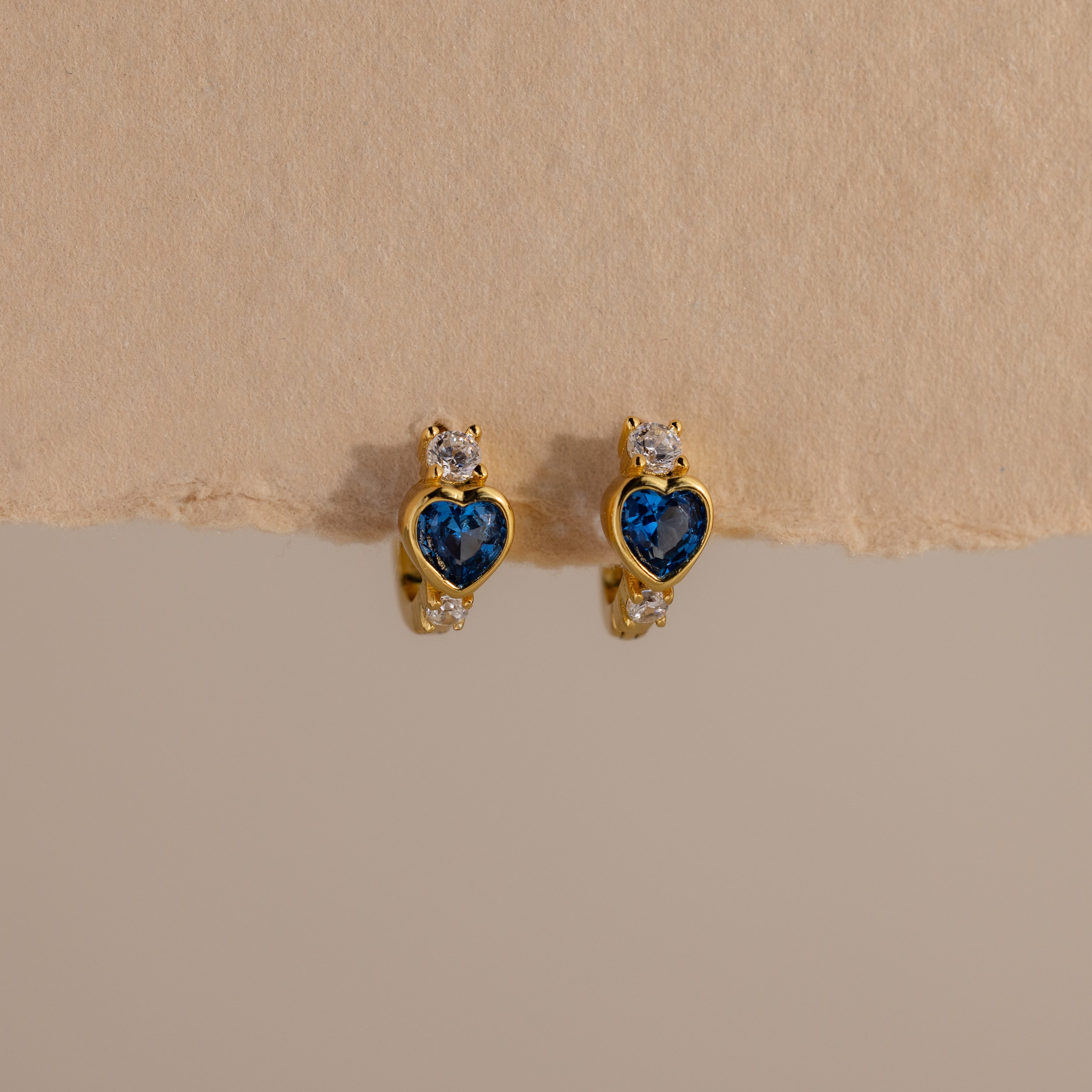 Blue Zircon Heart Hoops with sparkling clear stones are displayed on a beige textured background. These earrings add unique charm to any jewelry collection.