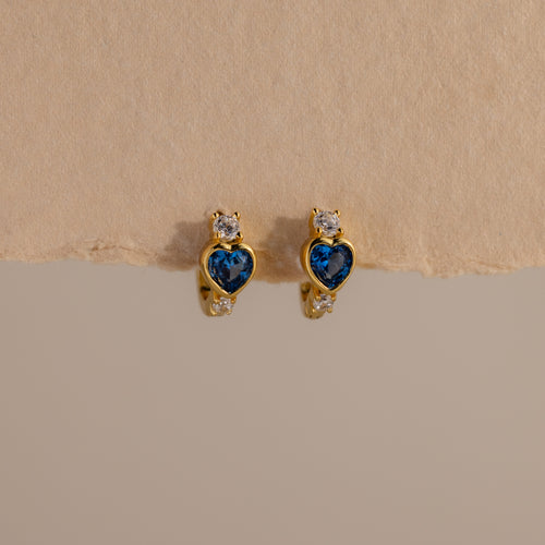 Blue Zircon Heart Hoops with sparkling clear stones are displayed on a beige textured background. These earrings add unique charm to any jewelry collection.
