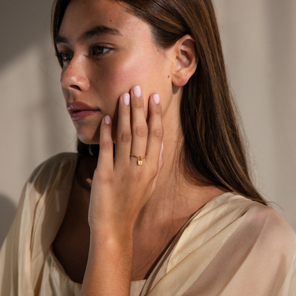 A woman with long brown hair touches her face, wearing the Initial Padlock Drop Ring and a sheer beige top—a chic example of personalized jewelry.
