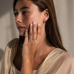A woman with long brown hair touches her face, wearing the Initial Padlock Drop Ring and a sheer beige top—a chic example of personalized jewelry.