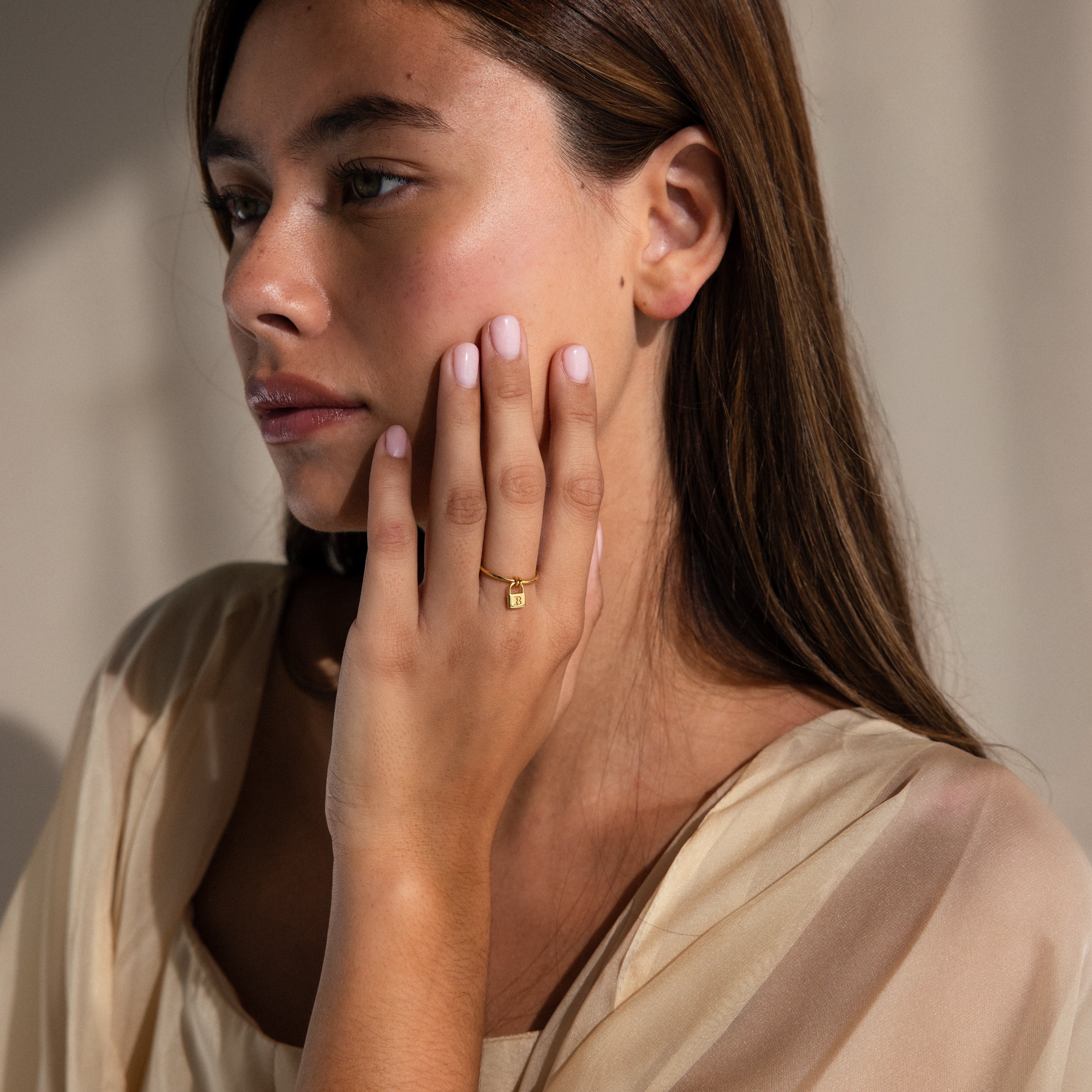 A woman with long brown hair touches her face, wearing the Initial Padlock Drop Ring and a sheer beige top—a chic example of personalized jewelry.
