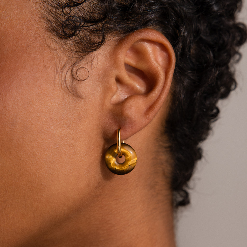 A person wears the Tiger's Eye Duo Hoops—round gold earrings with touches of Tiger's Eye gemstone—while their curly hair gently frames the sparkling accessory.