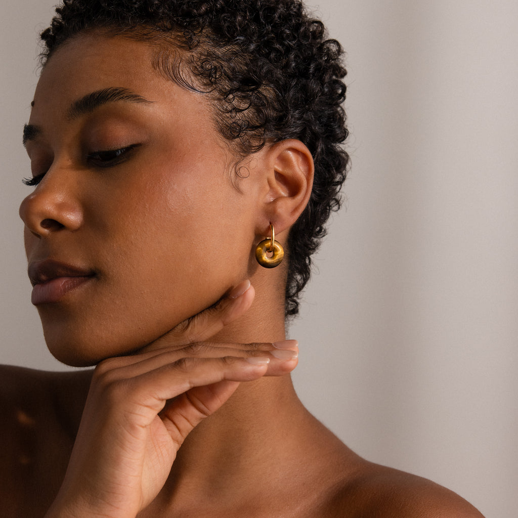 A woman with short curly hair wears Tiger's Eye Duo Hoops and touches her jaw with her hand.