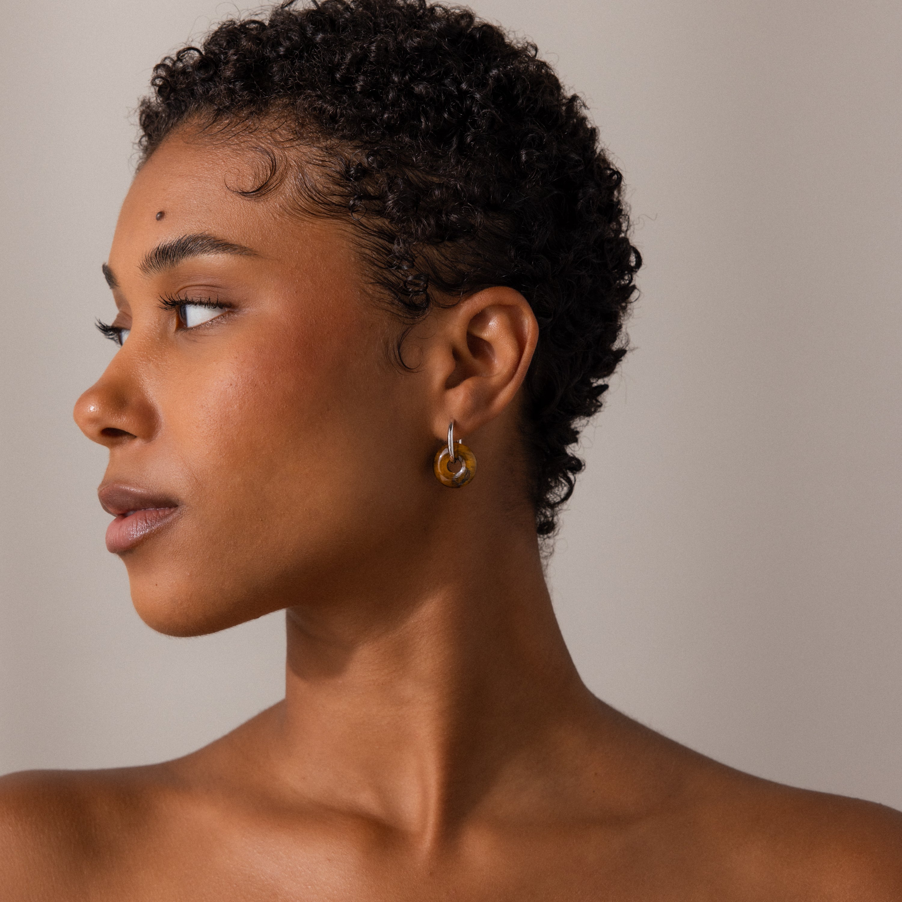 A woman with short curly hair wears Tiger's Eye Duo Hoops and looks to the left against a neutral background.