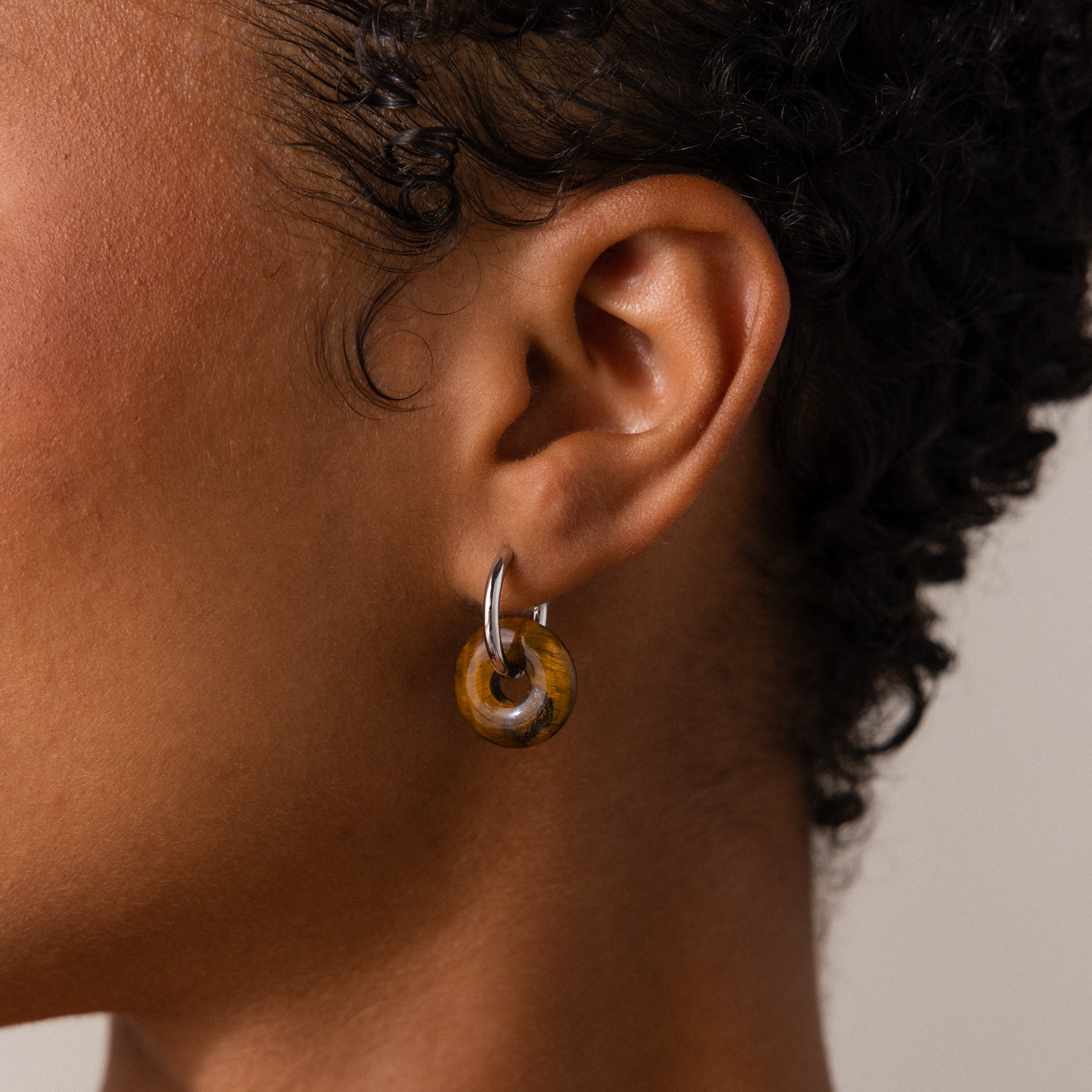 A close-up of a person wearing the Tiger's Eye Duo Hoops in White Gold, featuring a silver hoop and a round amber-like pendant that highlights the allure of golden-brown jewelry.