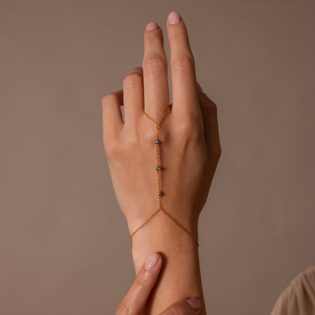 A hand wears the Turquoise Hand Chain Bracelet in 18K Gold, showcasing delicate chains and a touch of turquoise against a neutral background.
