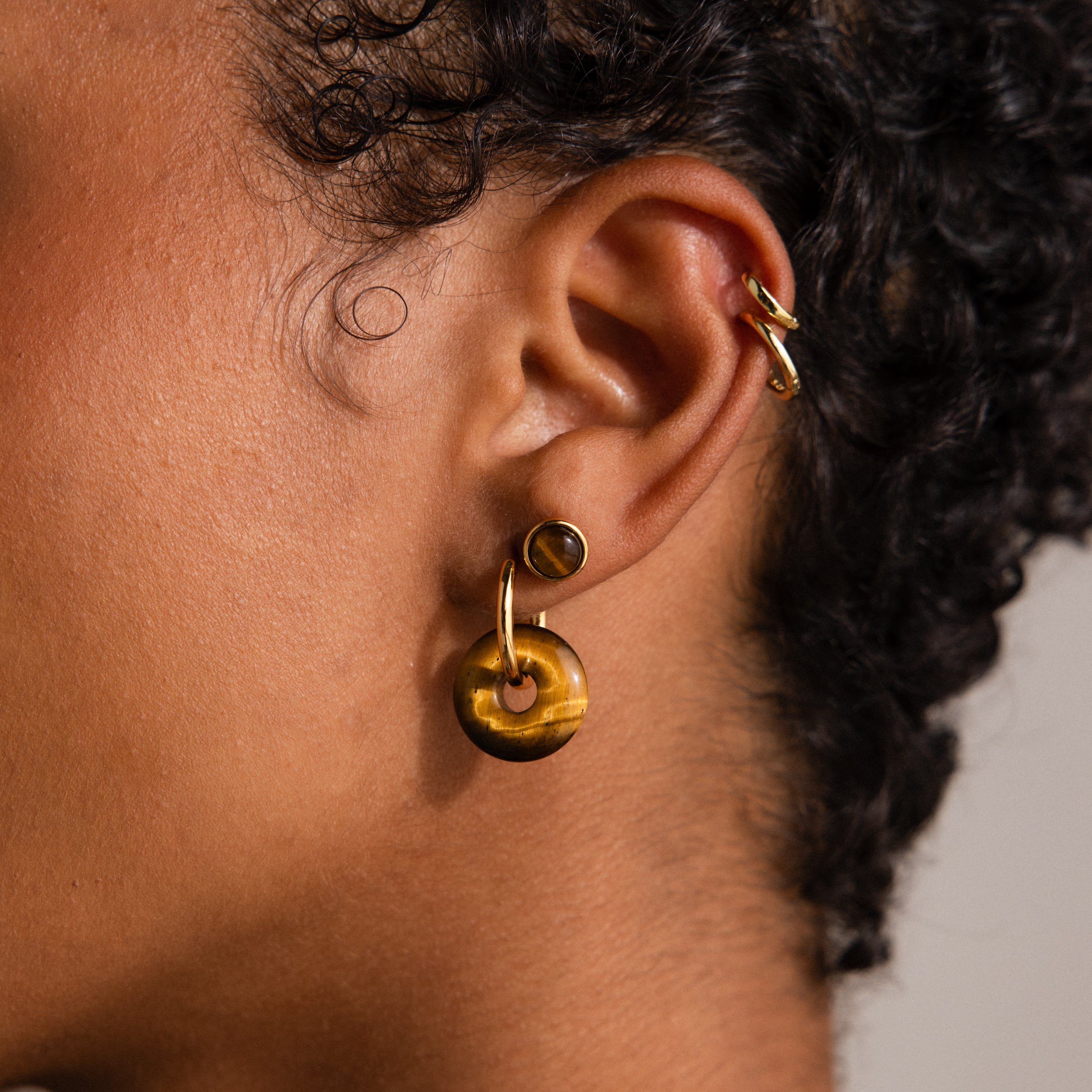 Close-up of an ear with curly hair adorned by Tiger's Eye Duo Hoops in 18K Gold, complemented by a stud and a gold ear cuff.