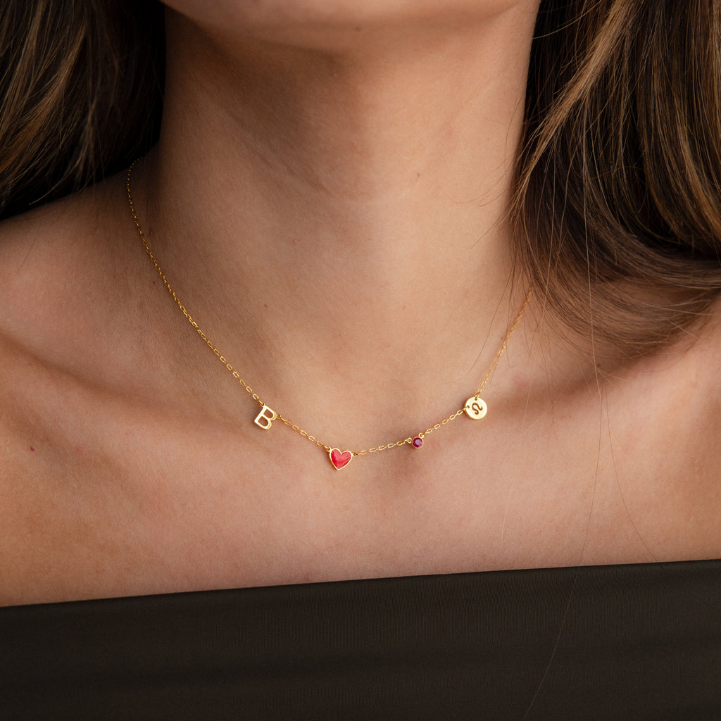 A woman wears the Dainty Space Charm Necklace in 18K Gold—a gold necklace featuring a B, red heart charm, pink gem, and gold smiley face—highlighting the charm of personalized jewelry.